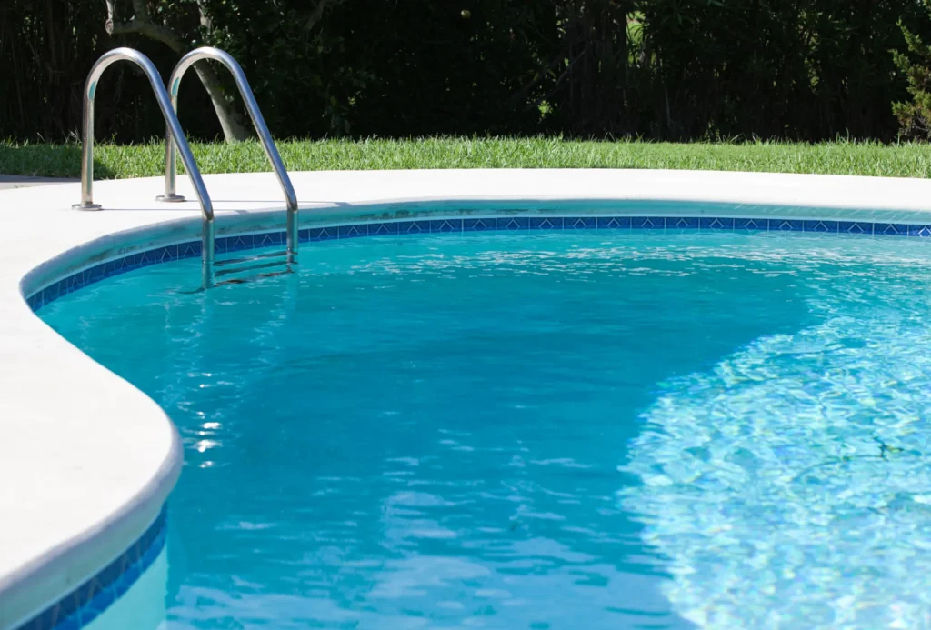 Close view of a pool corner with a stainless steel ladder and blue tile waterline next to a lawn.