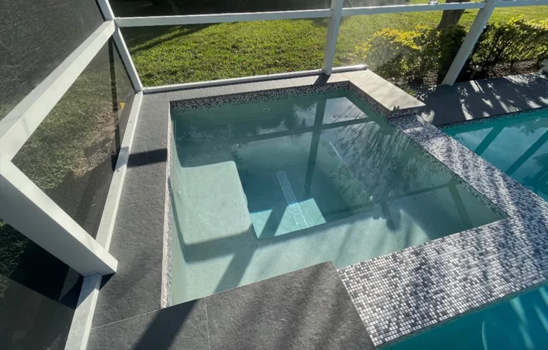 A raised spa with mosaic tile walls and stone coping sits beside a screened pool deck.