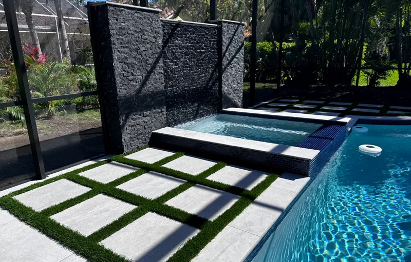 A dark stacked-stone water wall spills into a raised spa beside a pool with a checkerboard turf and paver deck under a screened enclosure.