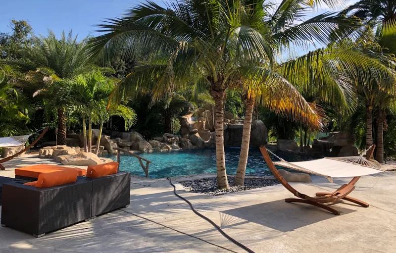A tropical lagoon-style pool with a rock waterfall surrounded by palm trees, hammocks, and lounge furniture.
