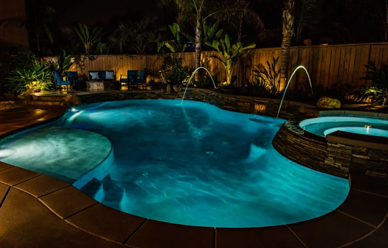 A freeform pool with a raised stone spa and two water arc jets lit at night in a backyard.