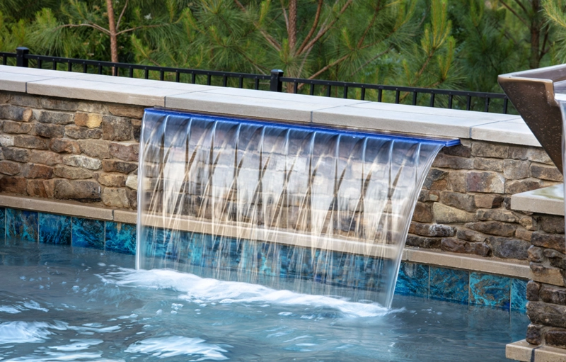 A wide sheer waterfall cascades over a stone wall into a pool with blue tile accents.