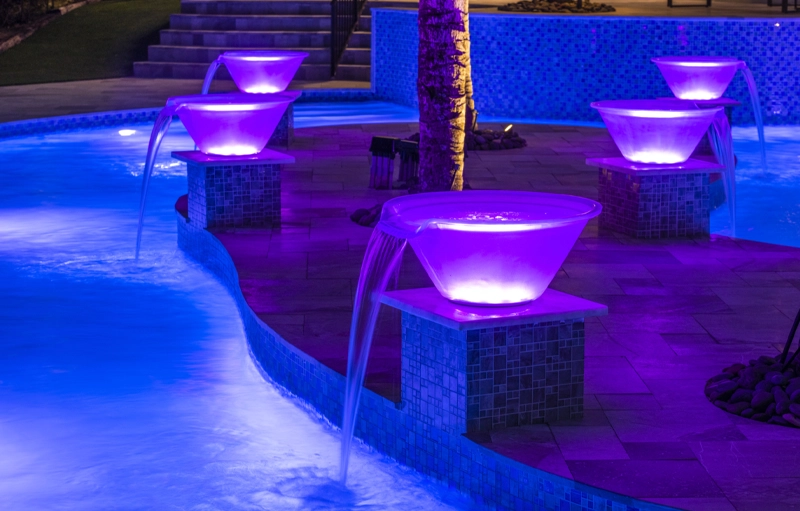 Five illuminated purple water bowls pour into a pool at night around a curved tiled edge.