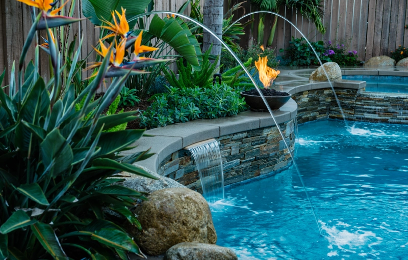A stone waterfall wall with a lit fire bowl and arcing water jets spills into a backyard pool at dusk.