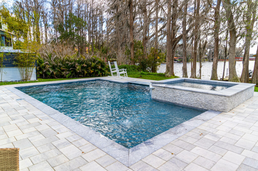 Rectangular pool with raised stone-clad spa on a paver deck beside a lake.