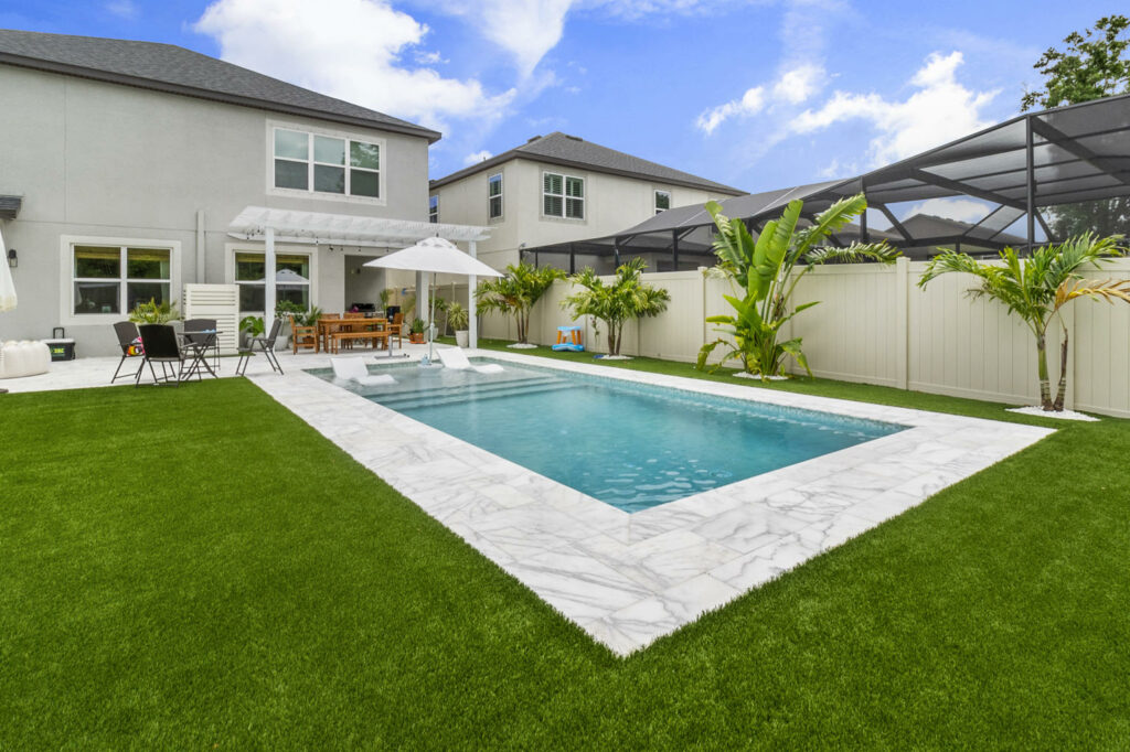 A rectangular backyard pool with a tanning ledge and loungers, surrounded by artificial turf and light stone pavers.