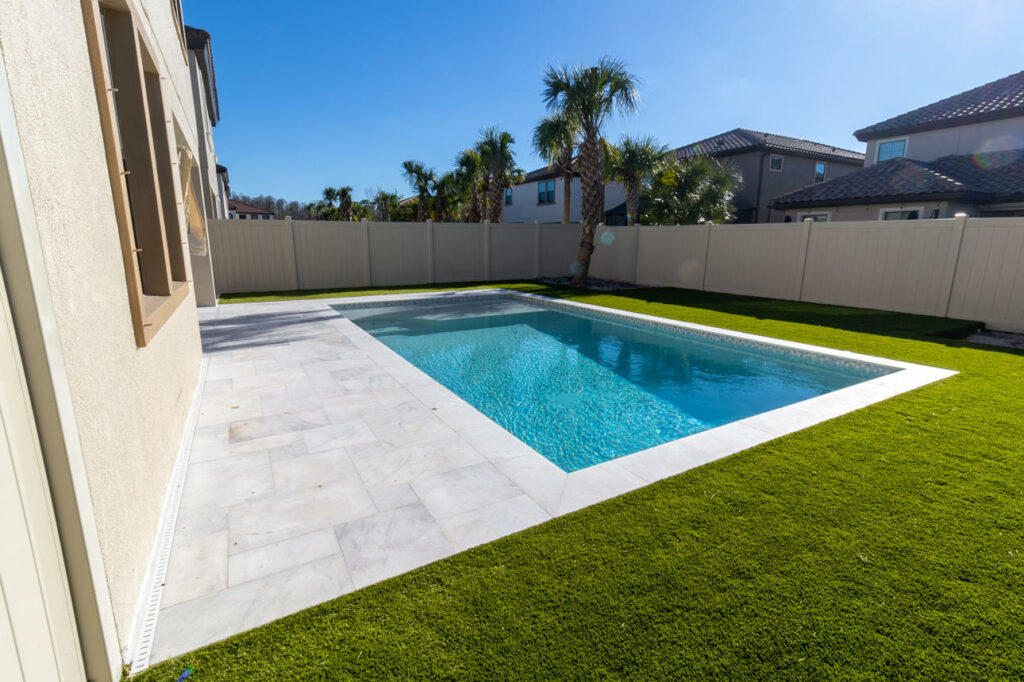 A small rectangular backyard pool with a paver deck and artificial turf surrounded by a vinyl privacy fence.