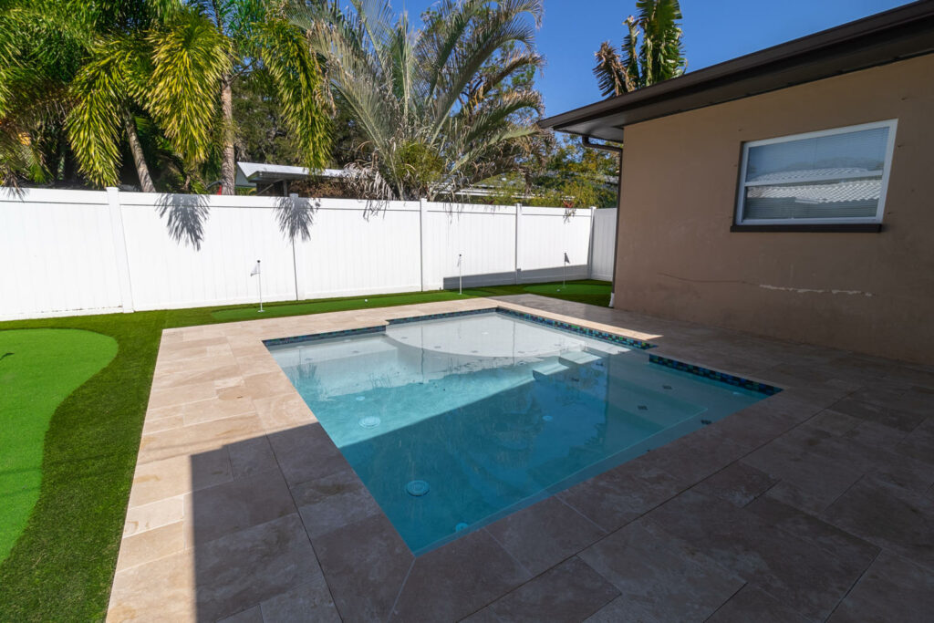 Small rectangular backyard pool with travertine deck and artificial turf putting green beside a white vinyl fence.