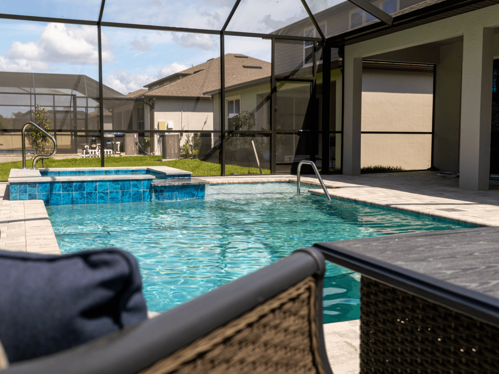 A screened backyard pool with a raised blue-tiled spa and travertine deck, patio furniture in foreground.