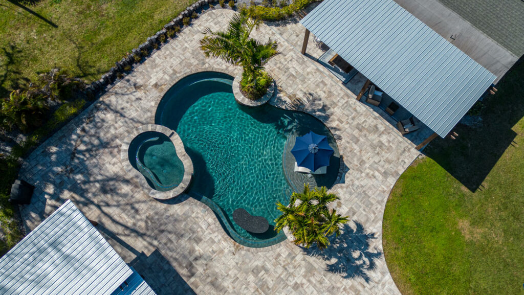 Aerial view of a freeform pool with an integrated spa and travertine paver patio surrounded by palm trees.