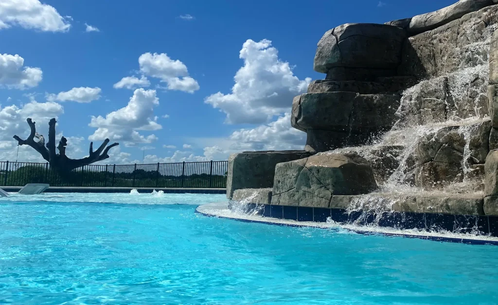 A large artificial rock waterfall cascades into a blue pool under a bright sky with scattered clouds.