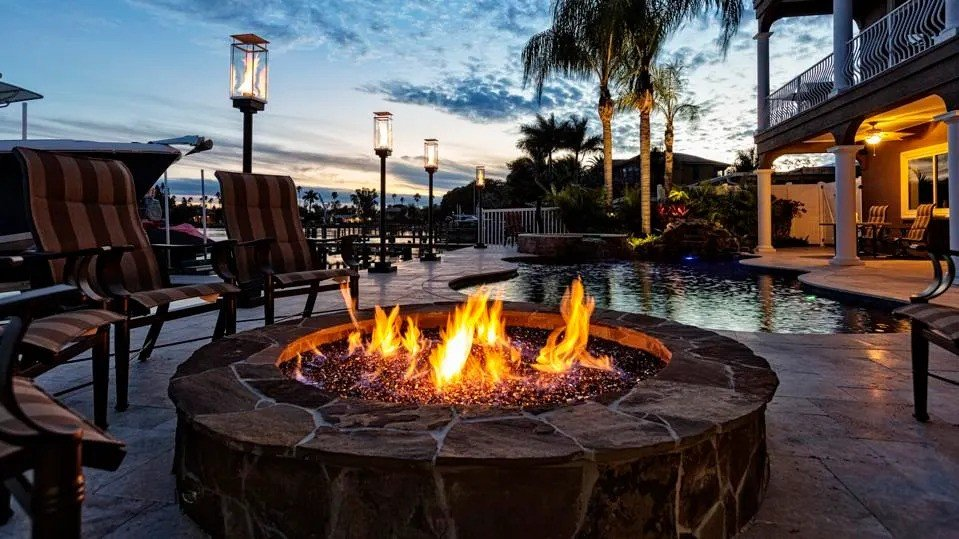 A large stone fire pit blazes on a waterfront patio with a pool and waterfall glowing behind it at dusk.