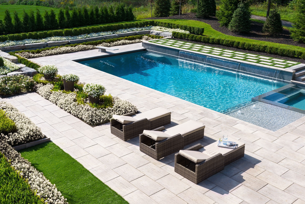 An aerial view of a rectangular backyard pool with a raised spillover spa, bordered by manicured hedges, flower beds, and a checkerboard turf accent, with lounge chairs on a stone patio.