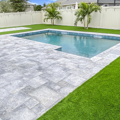 A small rectangular backyard pool with travertine pavers and artificial turf bordered by a tan privacy fence.