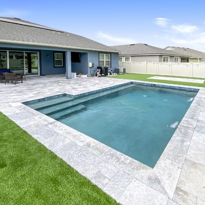 A small rectangular backyard pool with steps, surrounded by travertine pavers and turf.