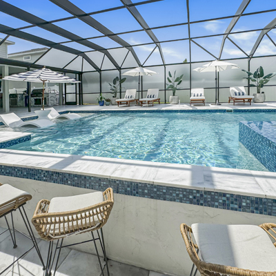 A rectangular pool with an integrated spa under a screened enclosure, surrounded by lounge chairs, umbrellas, and a bar seating area.