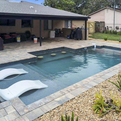 An L-shaped backyard pool with a paver deck and two white in-pool loungers on a shallow ledge.
