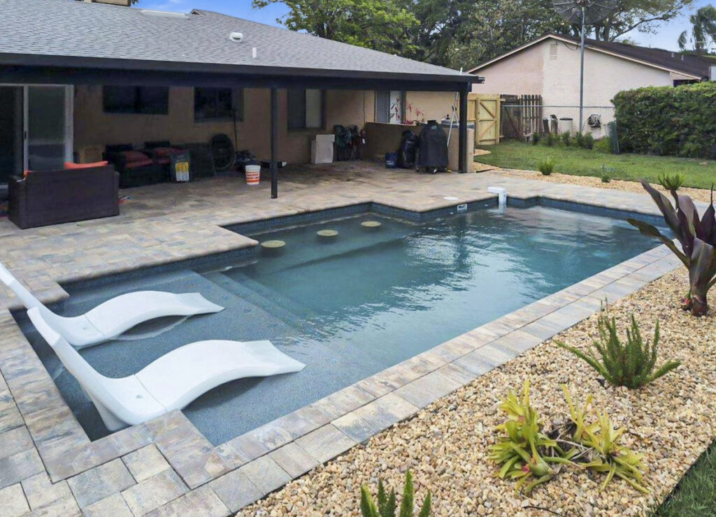A small rectangular backyard pool with a shallow ledge holding two white lounger chairs and a paver patio.