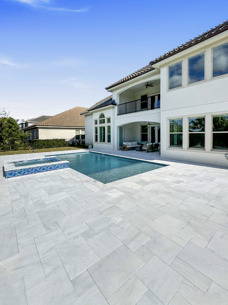 A rectangular backyard pool with a raised spillover spa sits beside a large stone paver patio behind a two-story Mediterranean-style home.
