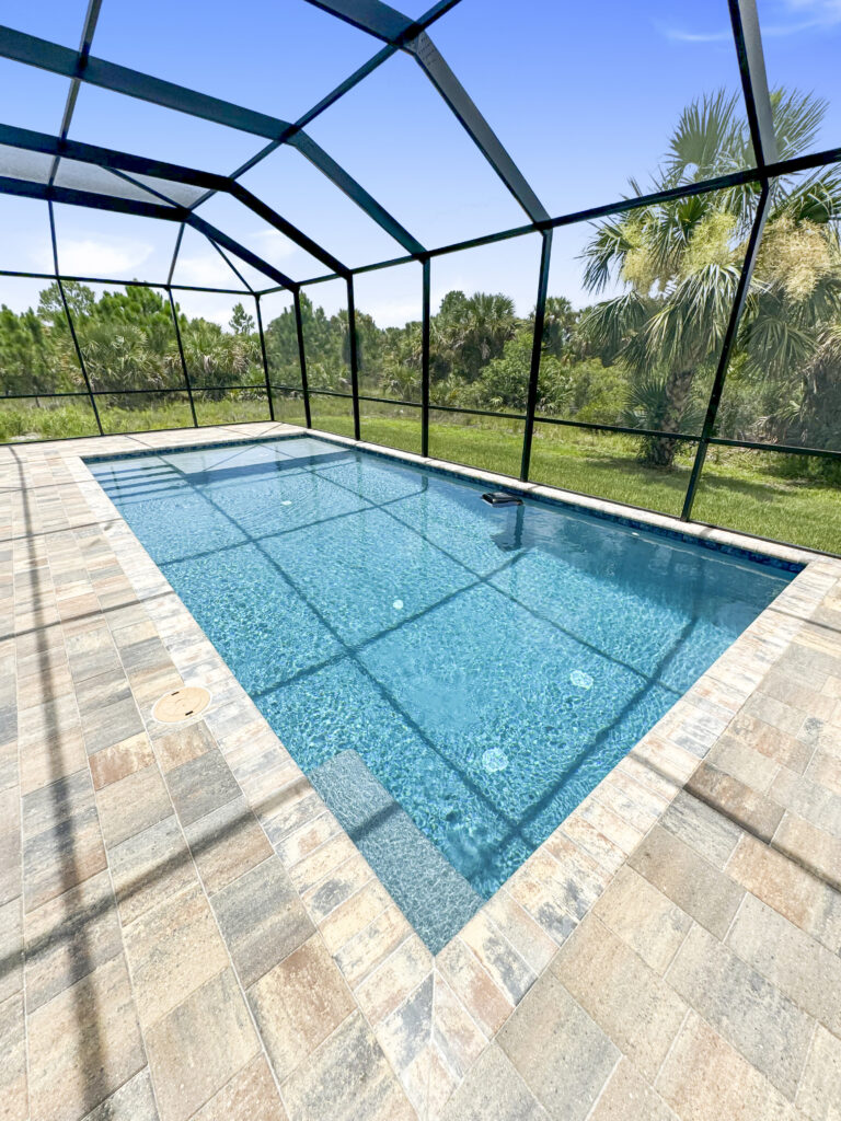 A screened rectangular pool with a shallow entry ledge and paver deck backing onto a wooded lot.