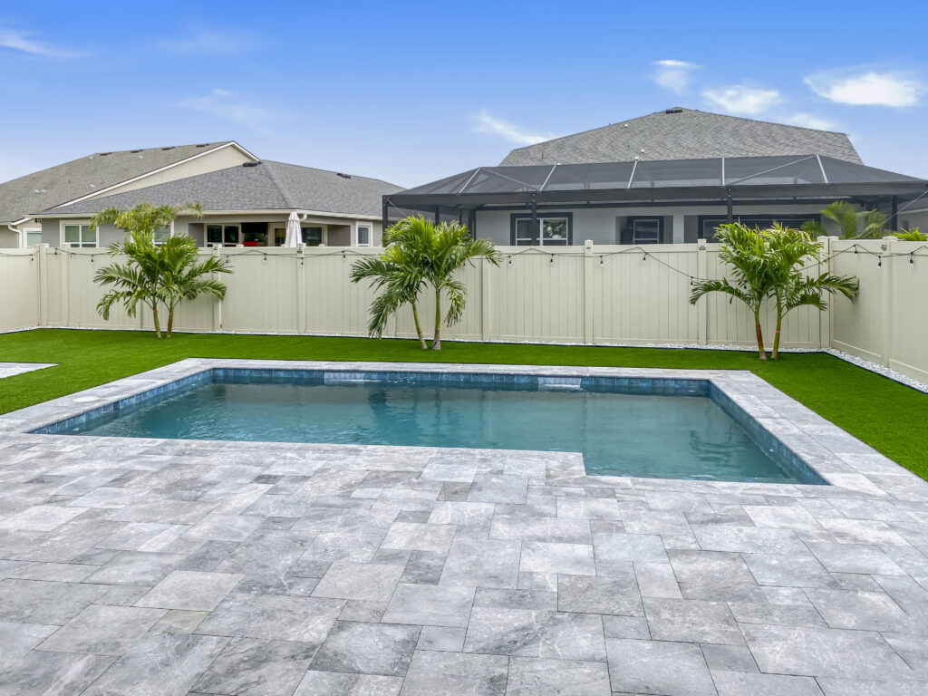 A small rectangular backyard pool with travertine decking and turf surrounded by a privacy fence and palm trees.