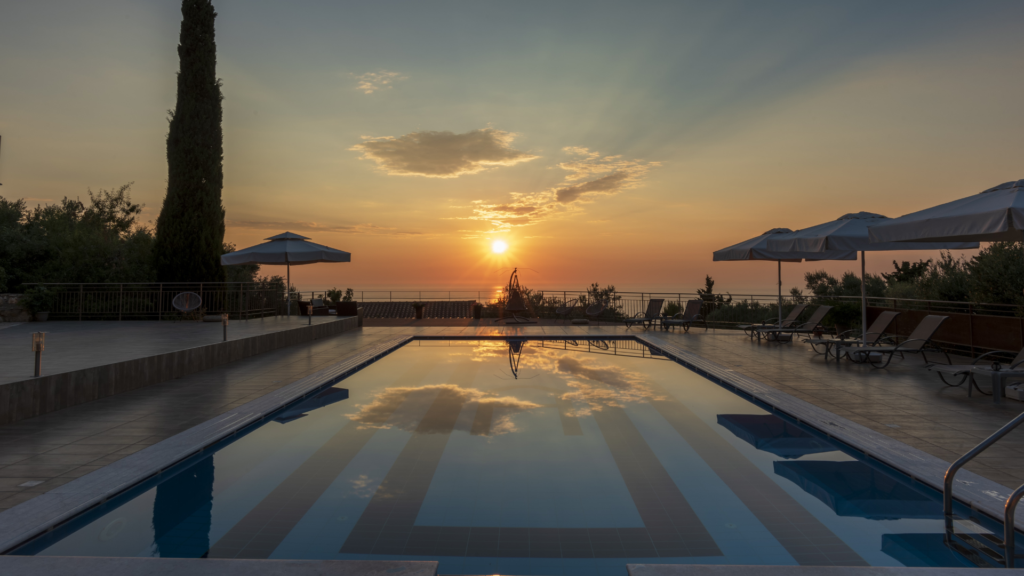 A rectangular resort pool reflecting a vivid sunset over the sea, flanked by loungers and umbrellas.