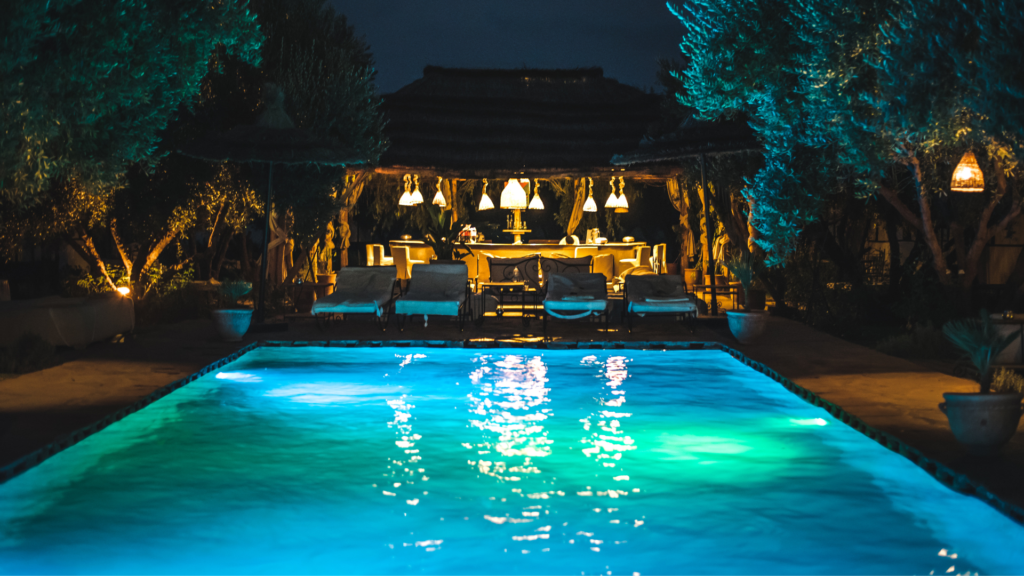 A rectangular pool glowing turquoise at night surrounded by trees and a lit lounge pavilion.