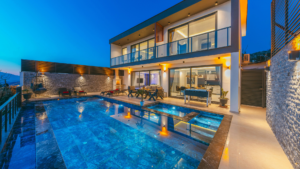 A modern two-story villa at dusk with an infinity-edge pool and raised spa lit by underwater and accent lighting.