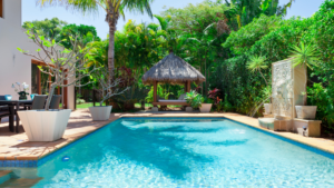 A rectangular backyard pool with a tiled waterline, bordered by a paver deck, tropical landscaping, a thatched-roof cabana, and a decorative stone fountain wall.
