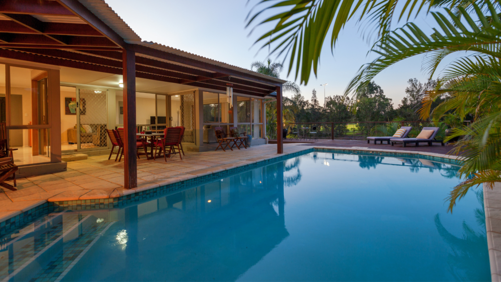 An L-shaped backyard pool at dusk beside a covered patio with loungers on the deck.