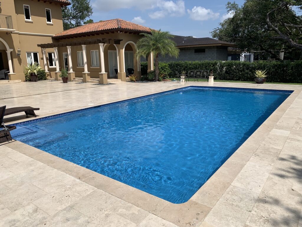 A rectangular pool with blue water and travertine deck in front of a Mediterranean-style home.
