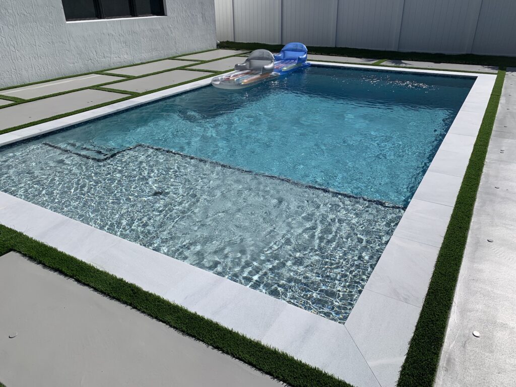 A small rectangular backyard pool with a shallow sun shelf and inflatable loungers, bordered by artificial turf and pavers.