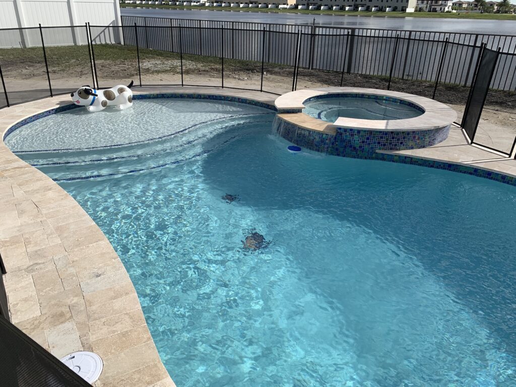A freeform pool with a raised spa and travertine deck overlooking a lake, with an inflatable dog float on the surface.