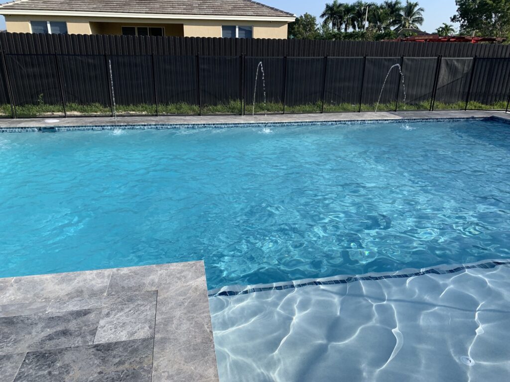 A rectangular backyard pool with three deck jets arcing water along one side, bordered by a dark privacy fence.