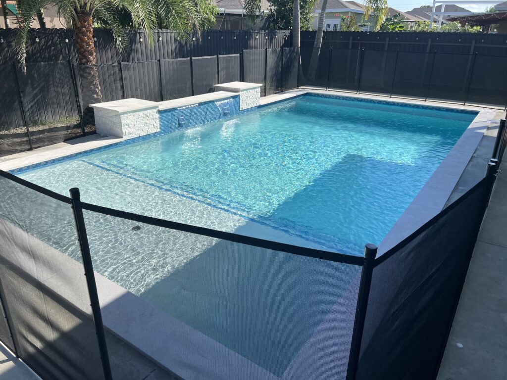 A small rectangular backyard pool with a raised stone water feature, enclosed by a black mesh safety fence.