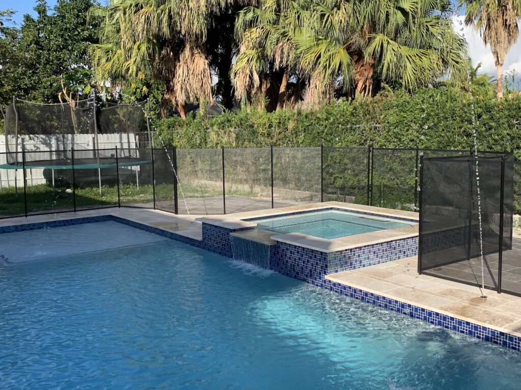 A raised spa with a spillway into a blue-tiled pool, enclosed by a black safety fence with a trampoline in the yard beyond.