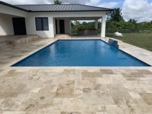 A rectangular pool with a small waterfall spillway sits on a travertine paver patio behind a modern house under open sky.