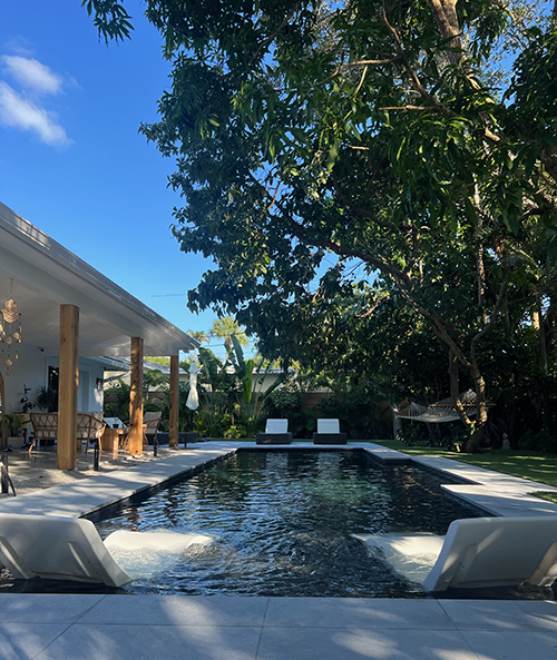 A dark-bottomed rectangular pool with two in-pool loungers sits beside a covered patio surrounded by lush trees.