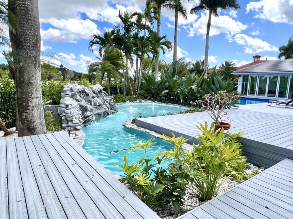 A lagoon-style pool with a rock waterfall grotto seen past a multi-level composite deck with tropical landscaping.
