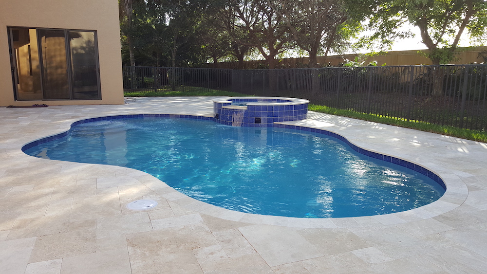 A freeform backyard pool with a raised blue-tiled spa spilling into the water, surrounded by travertine pavers and a metal fence.