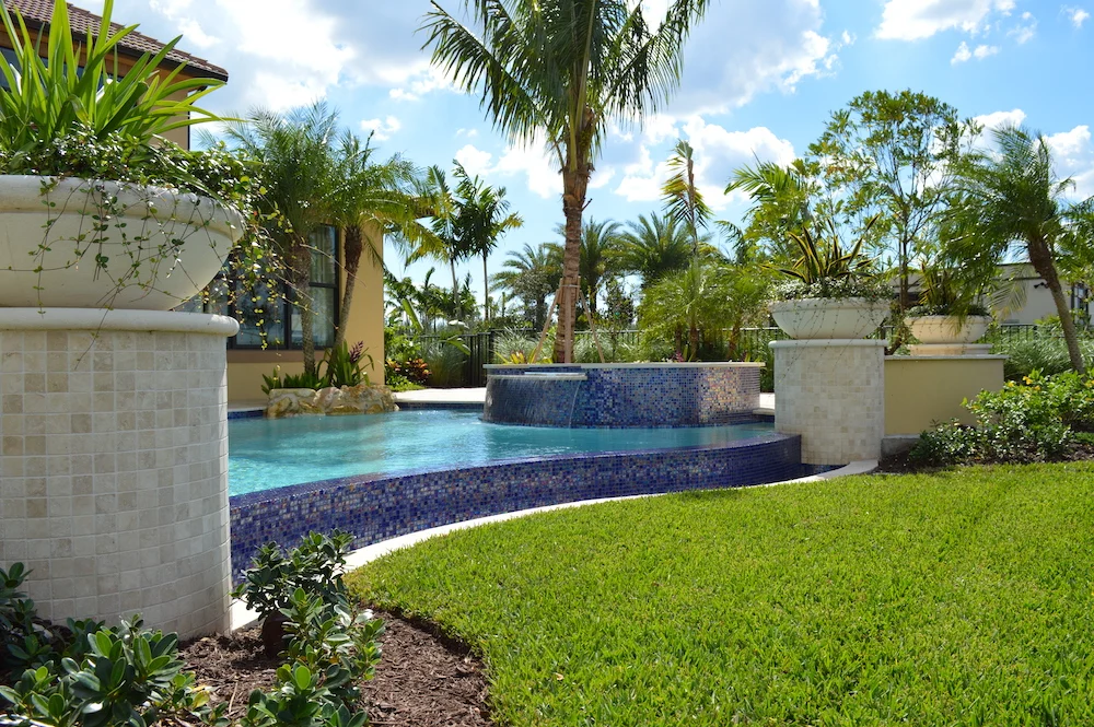 A raised spa with blue mosaic tile spills water into a pool edged by lush landscaping and palm trees.