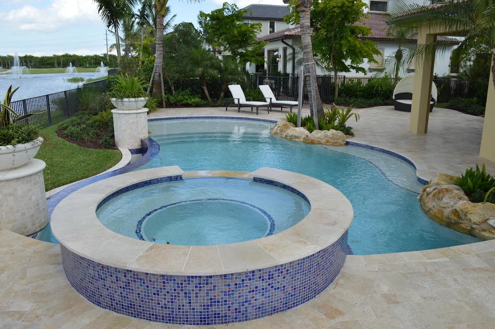 A raised round spa with blue mosaic tile fills the foreground with a freeform pool and lake view behind.