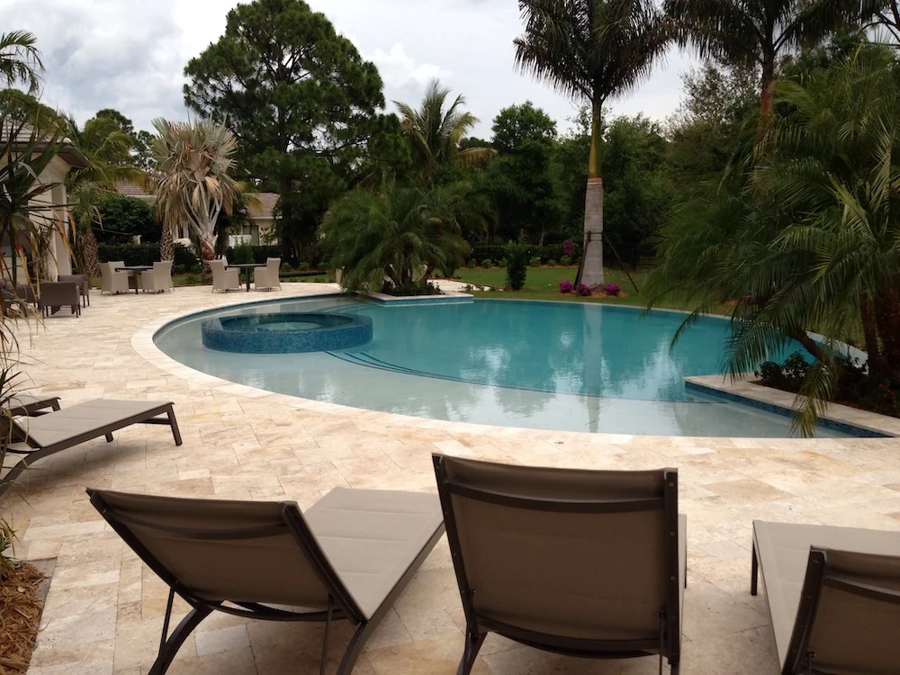Freeform backyard pool with a round raised spa surrounded by travertine deck and lounge chairs, framed by palm trees.