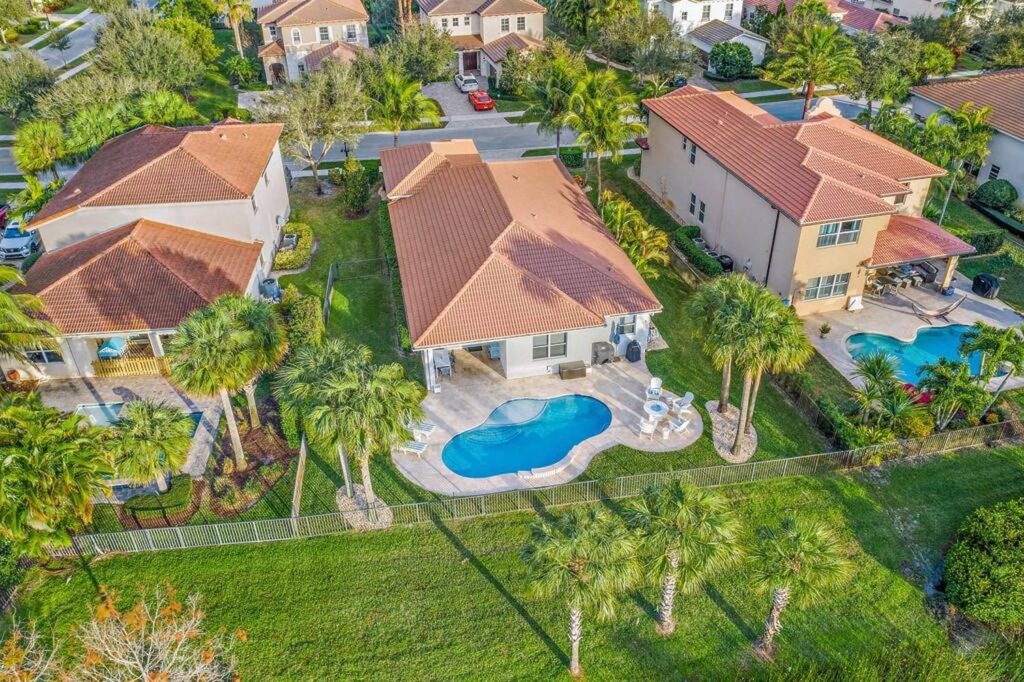 Aerial view of a small freeform pool behind a suburban house with palm trees and a fenced yard.
