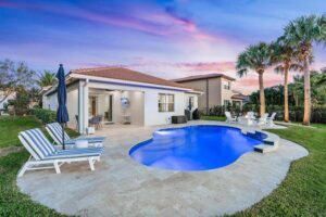 A small freeform pool with travertine deck behind a home at dusk with palm trees.