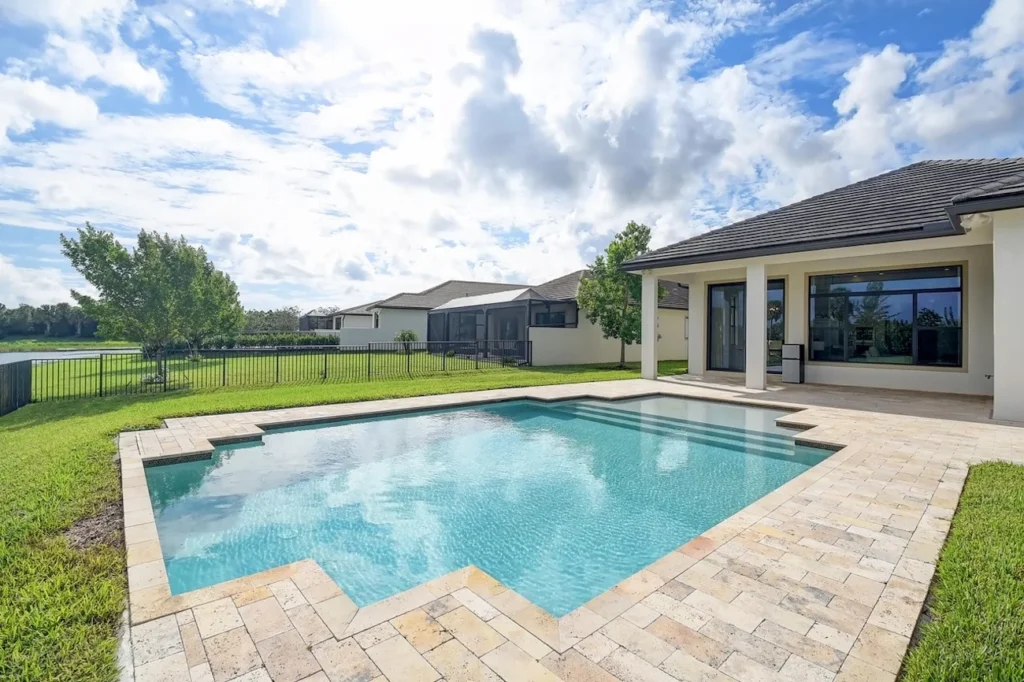 A geometric rectangular pool with notched corners on a travertine paver patio behind a modern single-story home with a pond and open lawn beyond.