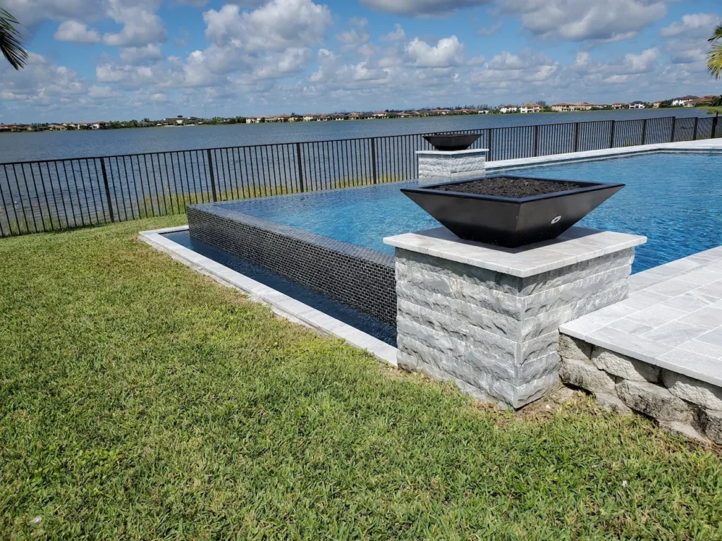 An infinity-edge pool corner with tiled spillway wall and two stone pedestals holding fire bowls overlooking a lake.