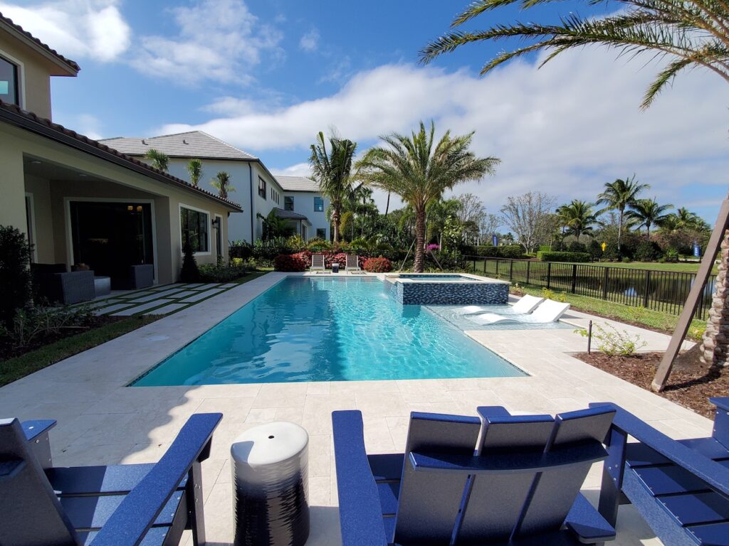 A rectangular backyard pool with a raised spa and in-pool loungers, set on a travertine patio with a lake view beyond the fence.
