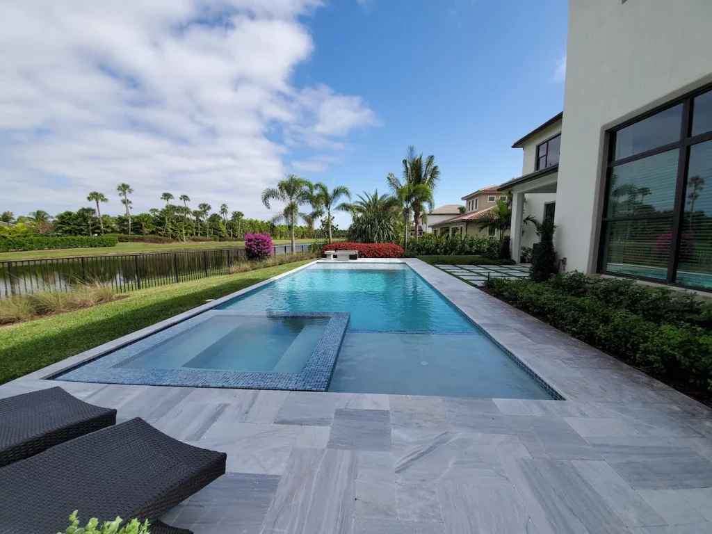 A rectangular gunite pool with an attached raised-edge spa sits on a stone patio behind a modern home overlooking a lake.