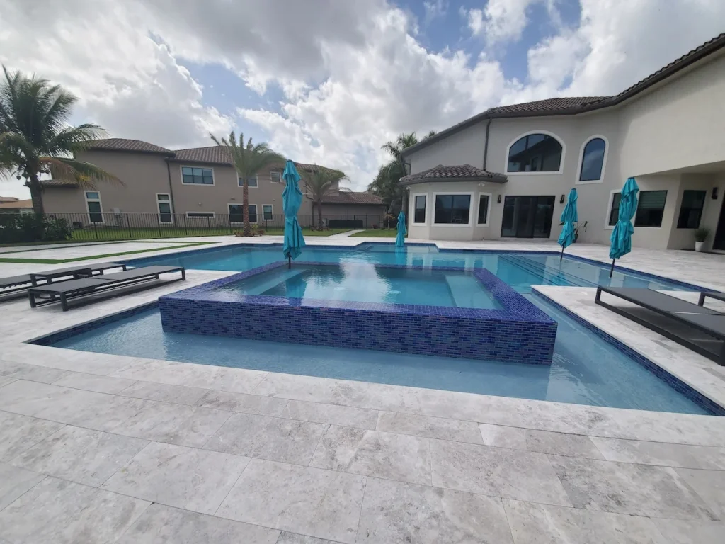 A large geometric pool with a raised blue-tiled spa in the center, surrounded by travertine pavers and a two-story stucco home.